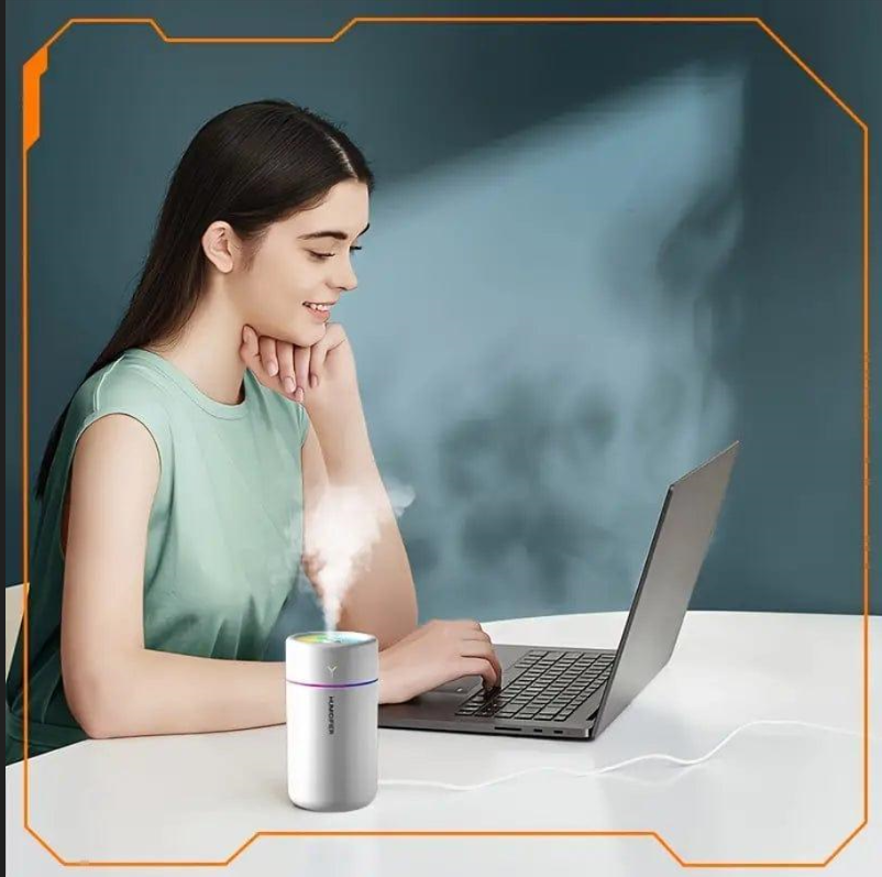 Woman using a laptop with a small device emitting steam on a table.